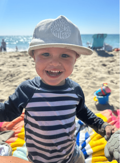 A young boy in a striped rash guard and hat is laughing at the beach.