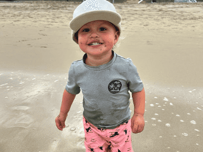 A young boy in printed swim shorts and a gray t shirt and hat wades in the ocean.