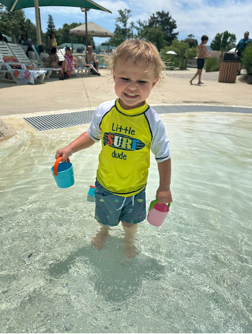 A young boy in a yellow rash guard with a surf board wades in a baby pool