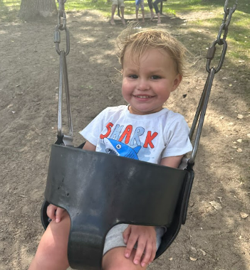 A blonde toddler in a shark t-shirt rides in a baby swing.